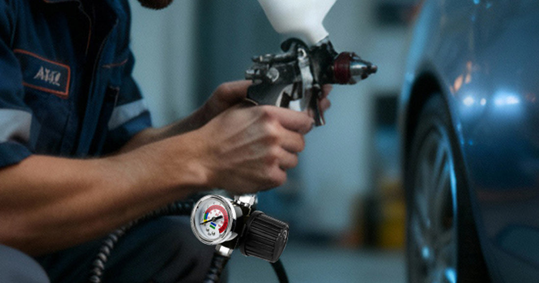 Mechanic using a spray gun with an inline air regulator for precise pressure control during car painting.